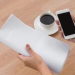 Mano sosteniendo un movil junto a una hoja de papel sobre la mesa